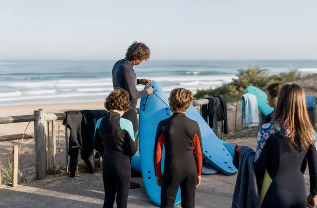 Anglet surf school group preparing softboards near dunes with waves behind them.