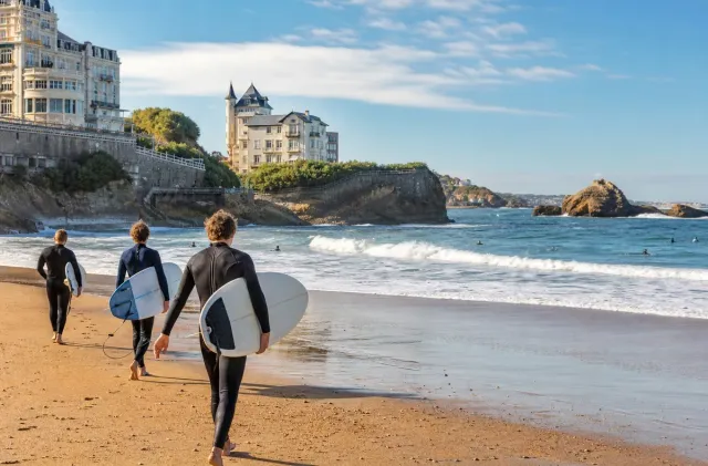Surfers carrying boards toward the water at a Biarritz beach below the Basque Coast cliffs.