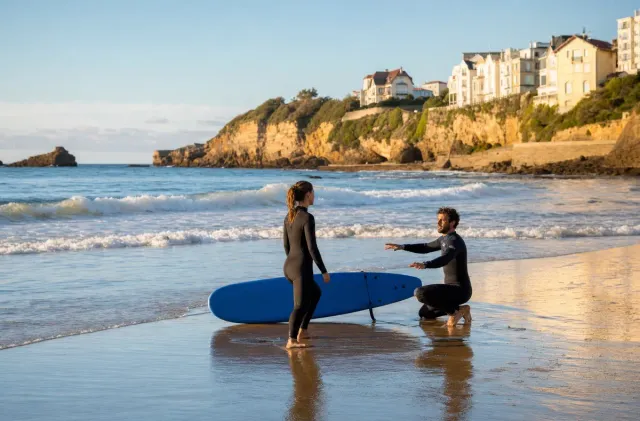 A surf instructor coaching a beginner with a softboard on wet sand at Cote des Basques in Biarritz.